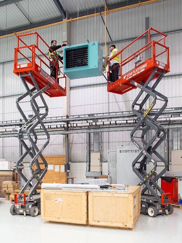 Two scissor lifts with men standing on the platforms fixing a machine that is suspended